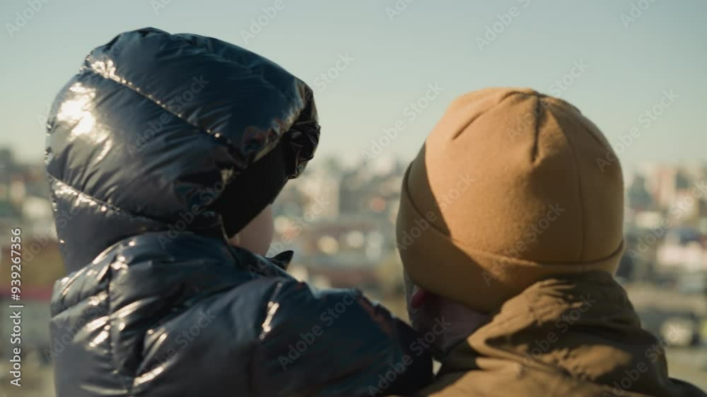 A father carrying his son looks out over a cityscape, while the son glances looks at the camera before turning away, both are dressed in jackets and beanies, with the blur cityscape