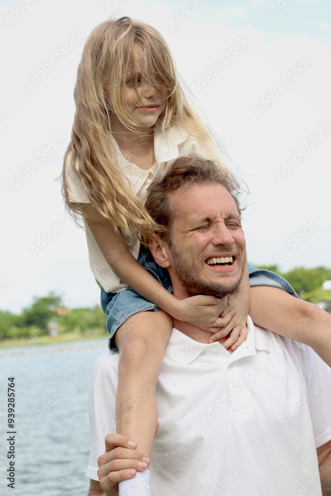 Obraz premium Father carrying cute daughter, joyful parent piggybacking his child girl at the park on summer holiday. Dad plays with kid and spends time outdoor together in beautiful garden. Happy family outside.