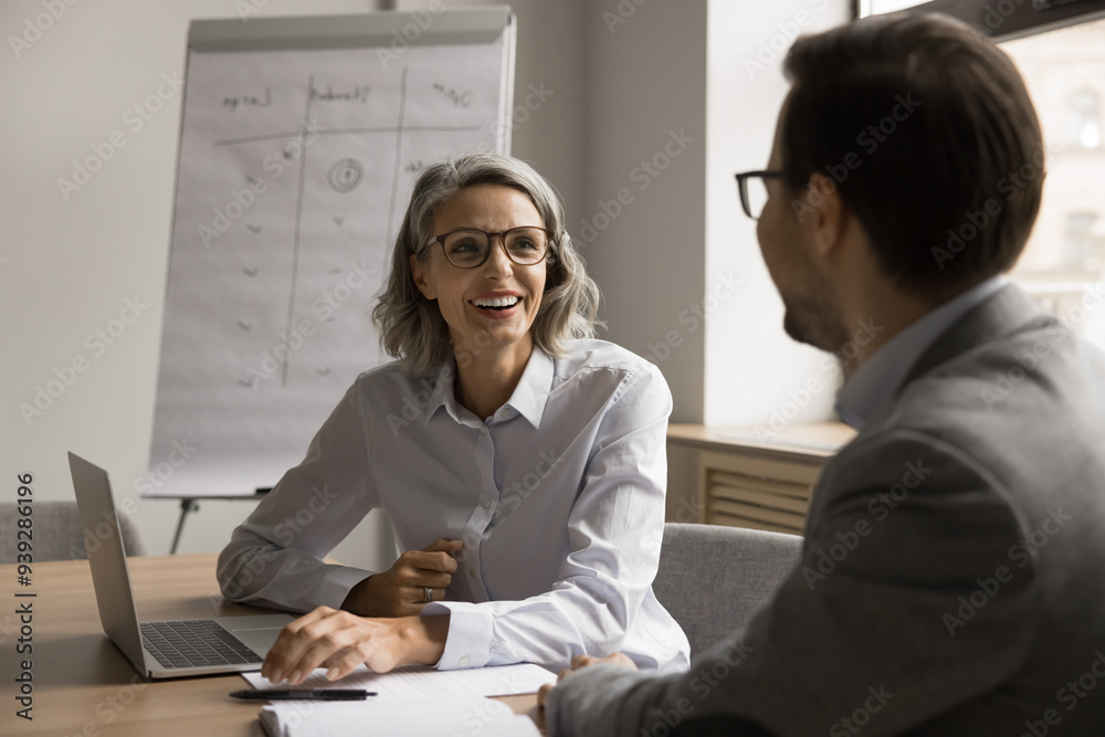 Fototapeta premium Happy mature woman negotiates with partner at conference table, lead pleasant business conversation, planning cooperation, working on task, selling company services to client during meeting in office