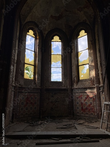 A dilapidated room featuring tall, stained glass windows with broken panes, scattered debris on the floor, and richly patterned walls in disrepair.