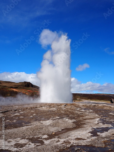 An impressive geyser eruption in Iceland captures a large plume of water and steam shooting into the clear blue sky, surrounded by the picturesque Icelandic landscape.