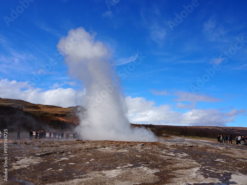 A geyser in Iceland erupts, propelling a powerful column of steam and water into the bright blue sky, surrounded by scenic and awe-inspiring landscape.