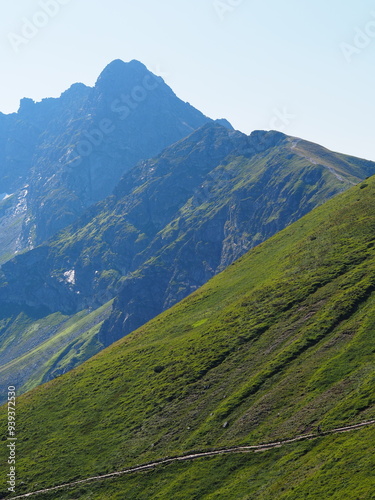 A majestic mountain peak rises above a green hillside, bathed in sunlight, highlighting the ruggedness and beauty of the natural landscape.