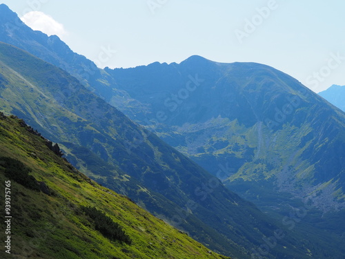 Fototapeta Naklejka Na Ścianę i Meble -  An expansive view of a mountain range with rocky peaks and lush greenery under a clear sky, showcasing the grandeur and unspoiled beauty of the natural environment.