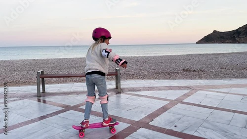 Blonde girl in helmet with safety features having fun practicing extreme sport skateboarding outdoors on the seaside promenade at sunset.