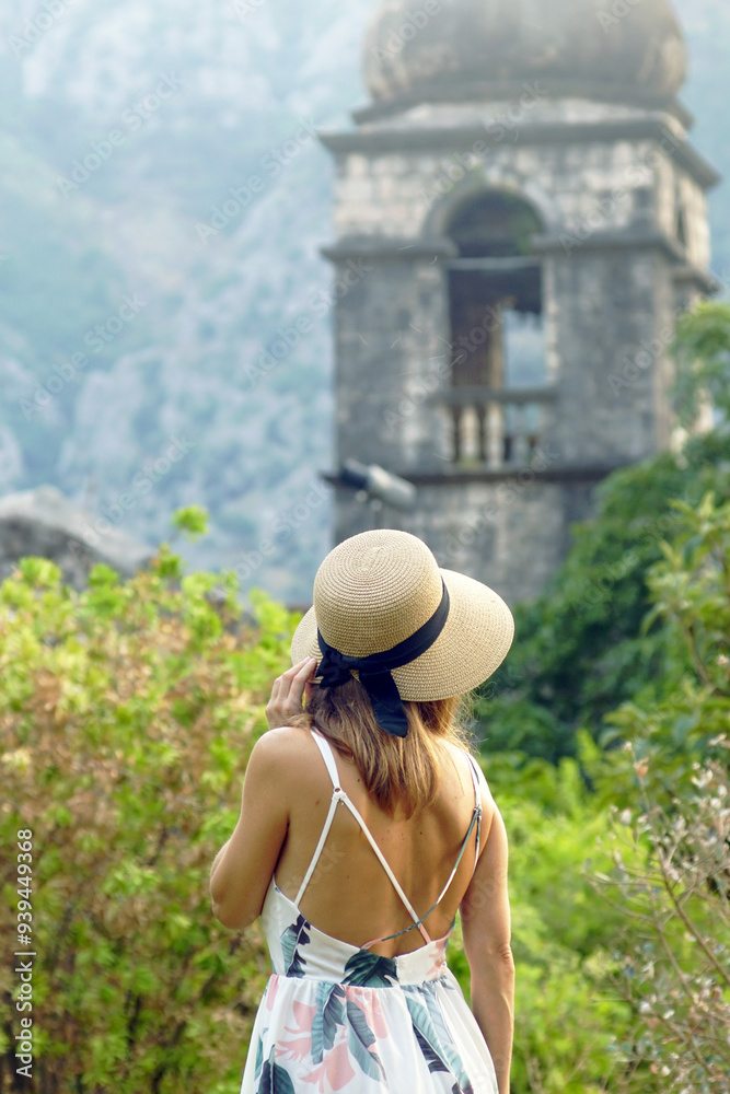 Fototapeta premium A female tourist sightseeing in the Old Town of Kotor: photographed from the back of a young woman in a hat, standing against the background of the bell tower of the Church of St. Clare (Montenegro)