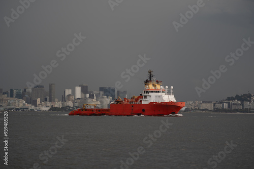 A bright red offshore supply vessel (OSV) navigates the calm waters near a city skyline, against a dark cloudy sky before storm. The ship's details, cranes, and deck equipment are clearly visible photo