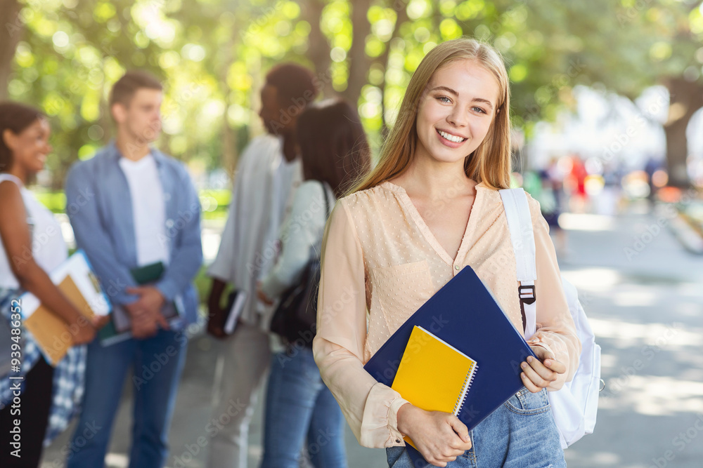 © Prostock-studio - Pretty smiling student girl with books posing at summer park, group of international chatting students on background © Prostock-studio - Pretty smiling student girl with books posing at summer park, group of international chatting students on background