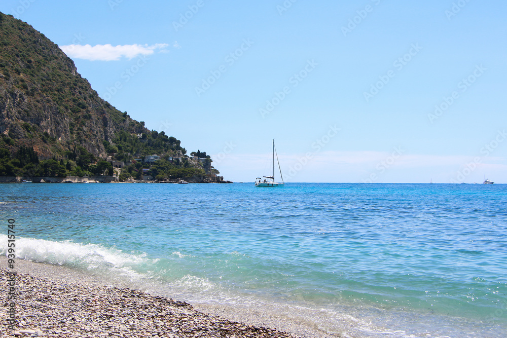 Pebble beach with gentle waves, turquoise sea, and a sailboat on the horizon. A rocky mountain rises to the left under a clear blue sky, creating a peaceful coastal view. Pebble beach with gentle waves, turquoise sea, and a sailboat on the horizon. A rocky mountain rises to the left under a clear blue sky, creating a peaceful coastal view.