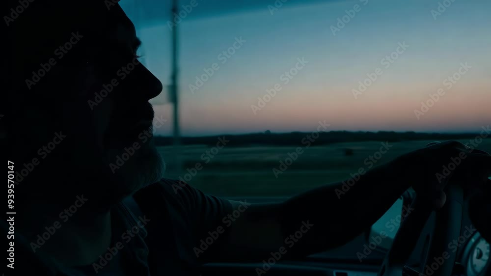 A man is seen driving through a serene landscape as the sun sets, deep in thought and enjoying the tranquility of the road. The soft hues of twilight create a reflective atmosphere.