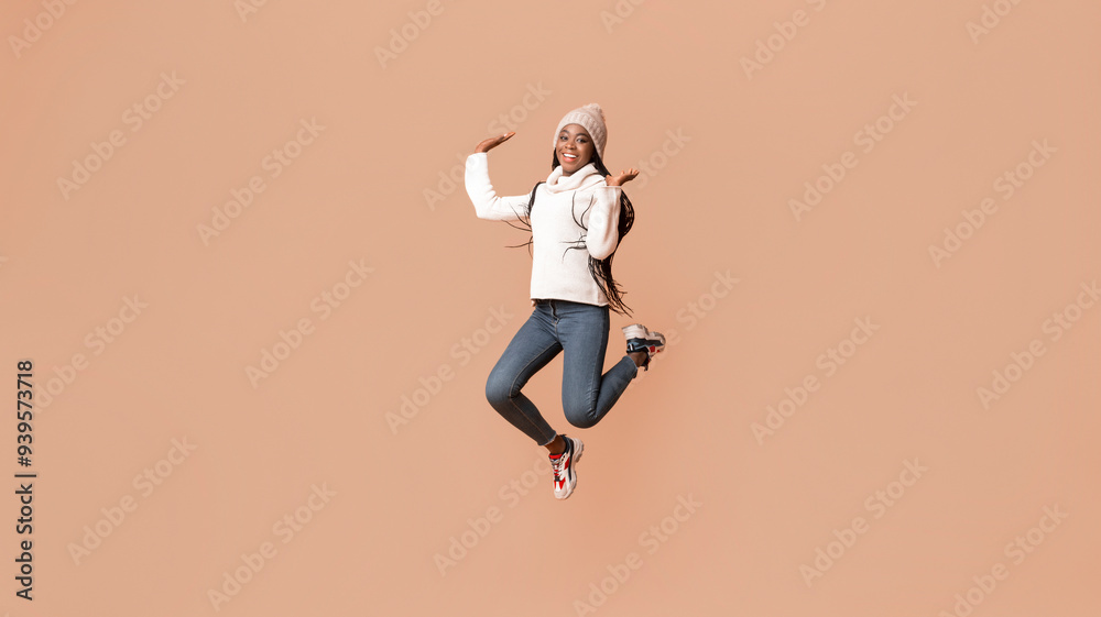 Fototapeta premium Playful young african woman in winter hat having fun over beige studio background, jumping in the air gracefully