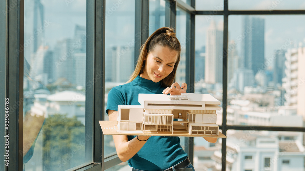 Young smart architect engineer holds architectural model while inspect house model. Professional interior designer checking house construction while standing near window with city view. Tracery