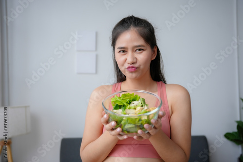 Canvas Print Young woman wearing sportswear is holding a bowl of salad and pouting because sh