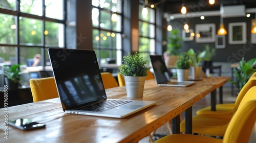 A modern, brightly lit co-working space with large windows, yellow chairs, and a wooden table adorned with laptops and small potted plants, creating an inviting work environment