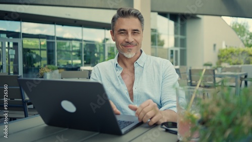 Smiling mature freelancer man working on laptop enjoying fresh air and freedom at restaurant terrace. Portrait of happy mid adult businessman. Male entrepreneur sitting at outdoor cafe.