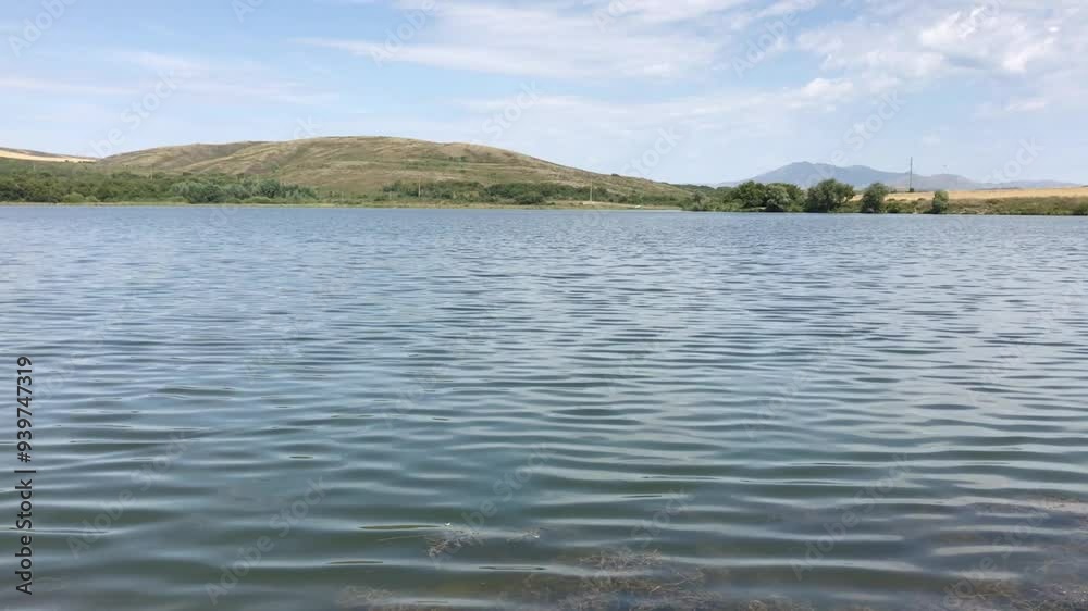 Serene Landscape of a Tranquil Lake with Rippling Water Under a Clear Blue Sky, Surrounded by Lush Greenery and Distant Rolling Hills, Capturing the Calmness of Nature on a Peaceful Summer Day Serene Landscape of a Tranquil Lake with Rippling Water Under a Clear Blue Sky, Surrounded by Lush Greenery and Distant Rolling Hills, Capturing the Calmness of Nature on a Peaceful Summer Day