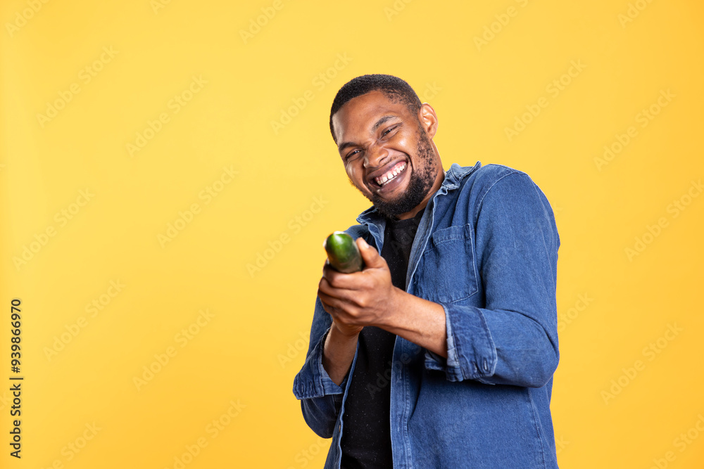 Young relaxed man pretending to shoot with cucumber as a gun, playing around and having fun with organic ripe veggie. Carefree funny person posing with raw food on yellow background. Young relaxed man pretending to shoot with cucumber as a gun, playing around and having fun with organic ripe veggie. Carefree funny person posing with raw food on yellow background.