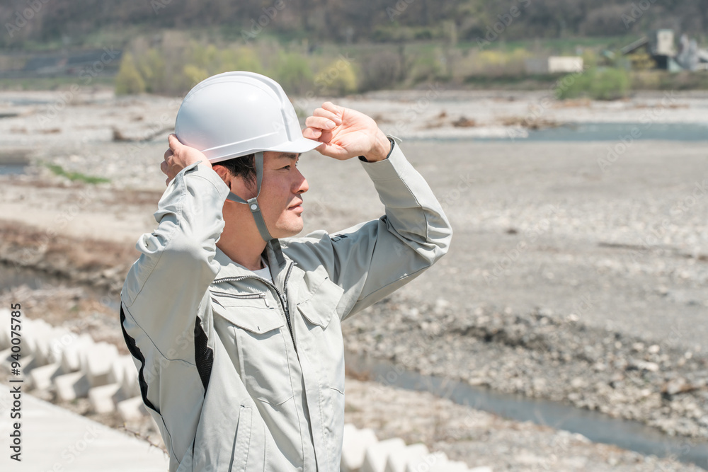 土木業・河川工事・建設現場に立つヘルメットを被った作業着姿のアジア人男性作業員
