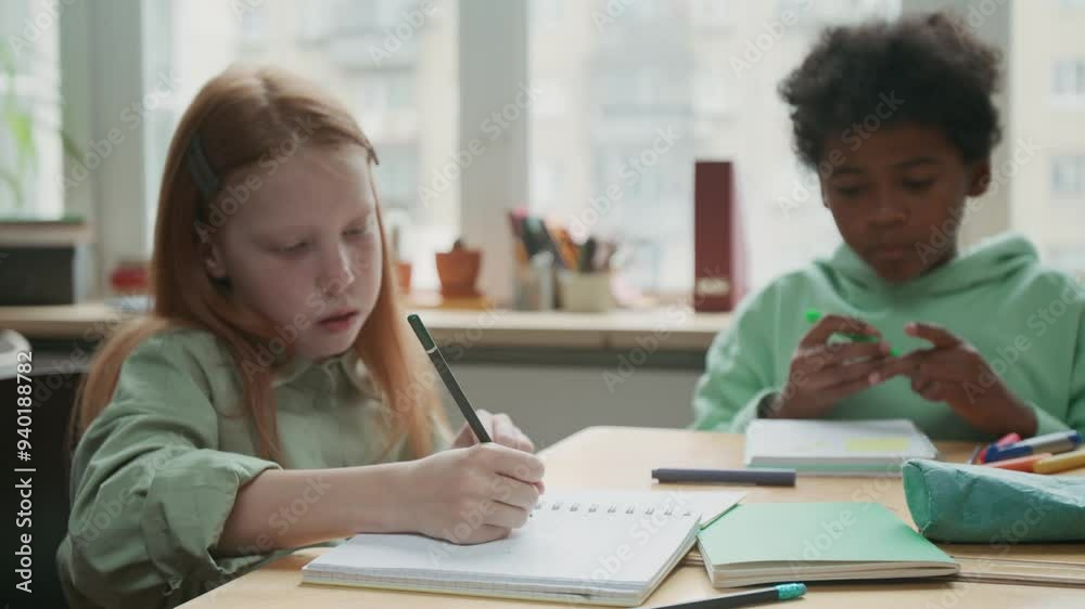 Medium shot of attentive, diligent 11-year-old Caucasian girl with long red hair and African American boy writing exercise or grammar rules in copybooks at English lesson Medium shot of attentive, diligent 11-year-old Caucasian girl with long red hair and African American boy writing exercise or grammar rules in copybooks at English lesson