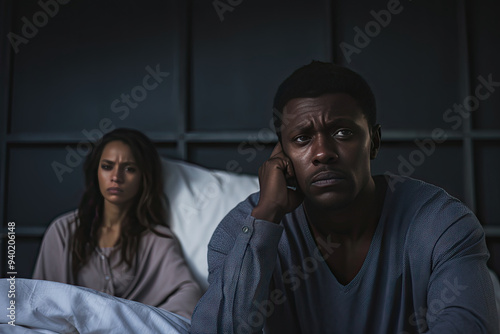 A tense moment between an African-American couple sitting in bed, with the man holding his head in worry and the woman looking concerned in the background, highlighting relationship struggles and emot photo