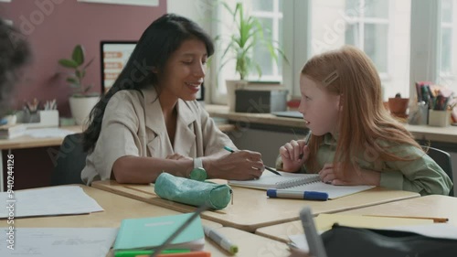 Medium zoom-in shot of multiethnic boy and girl writing exercise in copybooks during English language lesson, while Hispanic female teacher is explaining rules to red-haired Caucasian schoolgirl photo