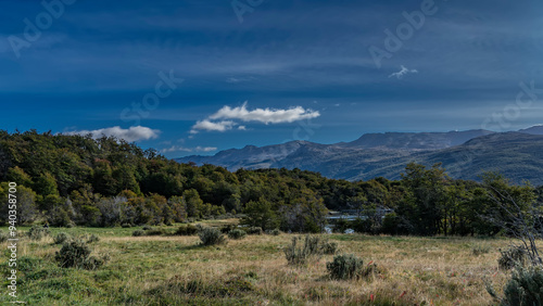 Beautiful autumn landscape. The yellowed grass in the meadow. A green forest on the shore of the lake. Picturesque mountains against a blue sky and clouds. Argentina. Tierra del Fuego National Park. 