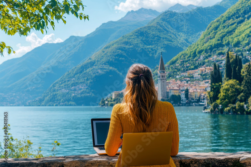 Happy female digital nomad freelancer working remotely by blue lake como in italy. Travelling woman on laptop outdoors with scenic view.