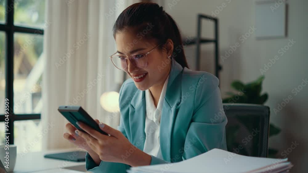 Businesswoman is calculating her business expenses using a calculator app on her smartphone. She is sitting at her desk in her office