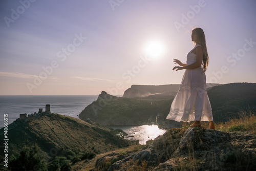 A woman stands on a rocky hill overlooking the ocean. She is wearing a white dress and she is enjoying the view. The scene is serene and peaceful, with the sun shining brightly in the background.