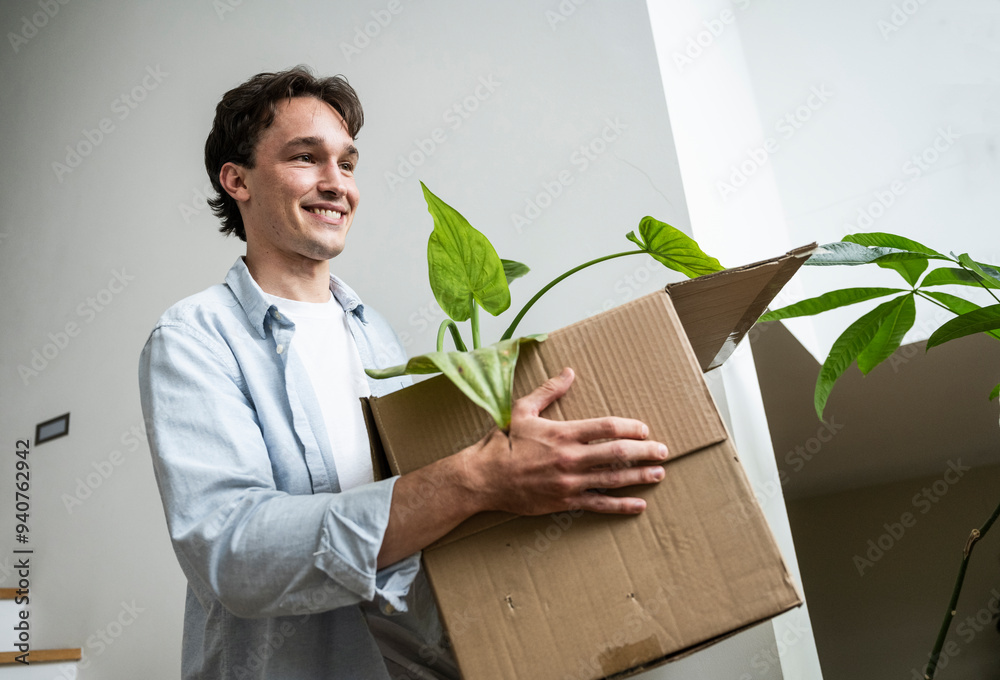 Smiling young man carrying cardboard box with houseplant near wall Smiling young man carrying cardboard box with houseplant near wall