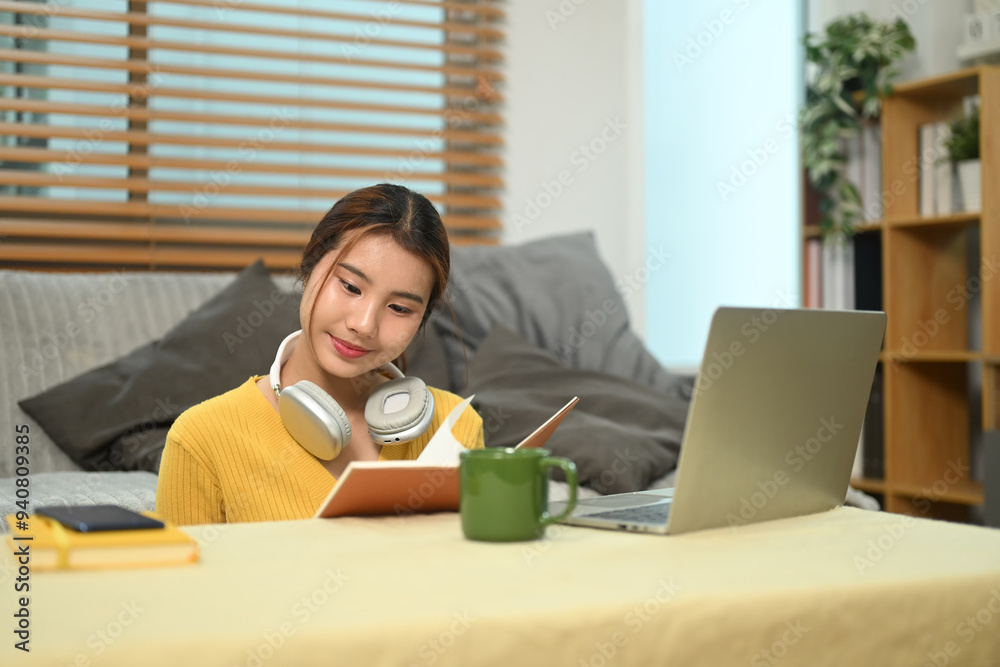 Smiling young asian girl sitting in living room using laptop and writing in a notebook Smiling young asian girl sitting in living room using laptop and writing in a notebook