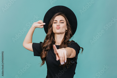 playful young woman wearing a black hat and black shirt, making a pouty face while pointing directly at the camera against a turquoise background photo