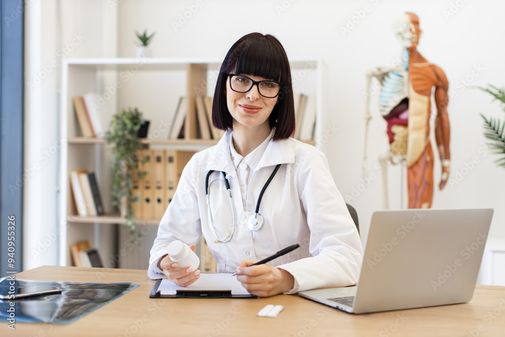 Smiling female doctor holding medication while working at desk in modern office with medical equipment and laptop Smiling female doctor holding medication while working at desk in modern office with medical equipment and laptop