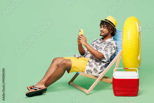 Wall Mural Full body side view young happy Indian man wear beach shirt hat rest near hotel pool sit in deckchair hold use mobile cell phone isolated on plain green color background