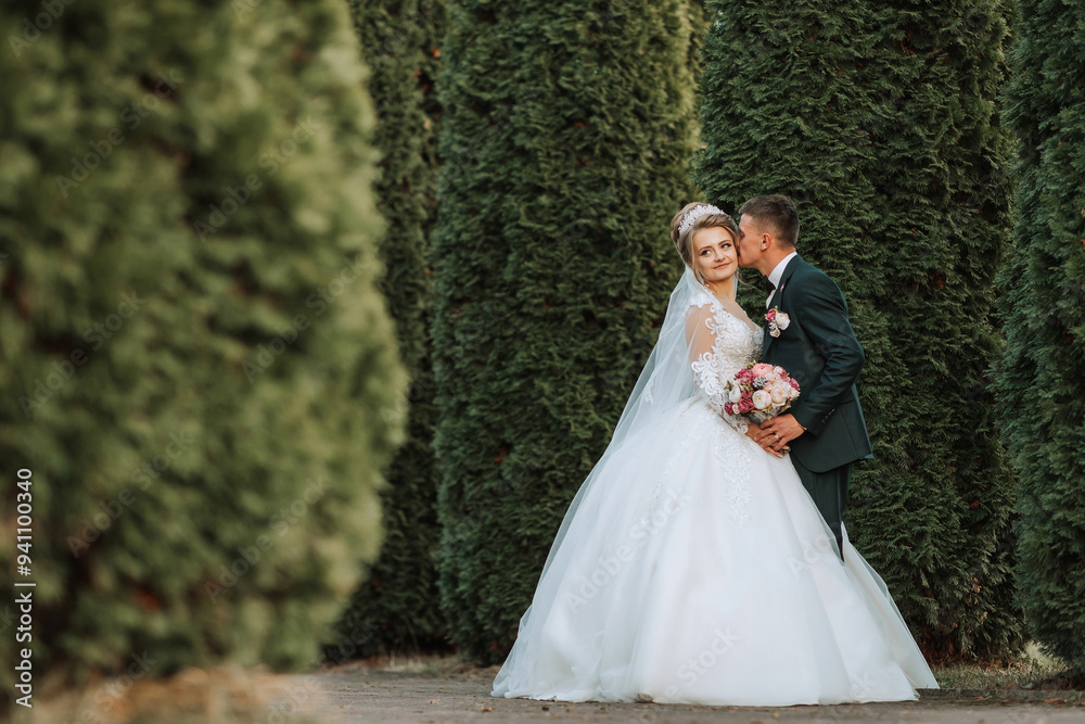 A bride and groom are posing for a picture in front of a hedge. The bride is wearing a white dress and the groom is wearing a suit. They are both smiling and looking at the camera A bride and groom are posing for a picture in front of a hedge. The bride is wearing a white dress and the groom is wearing a suit. They are both smiling and looking at the camera