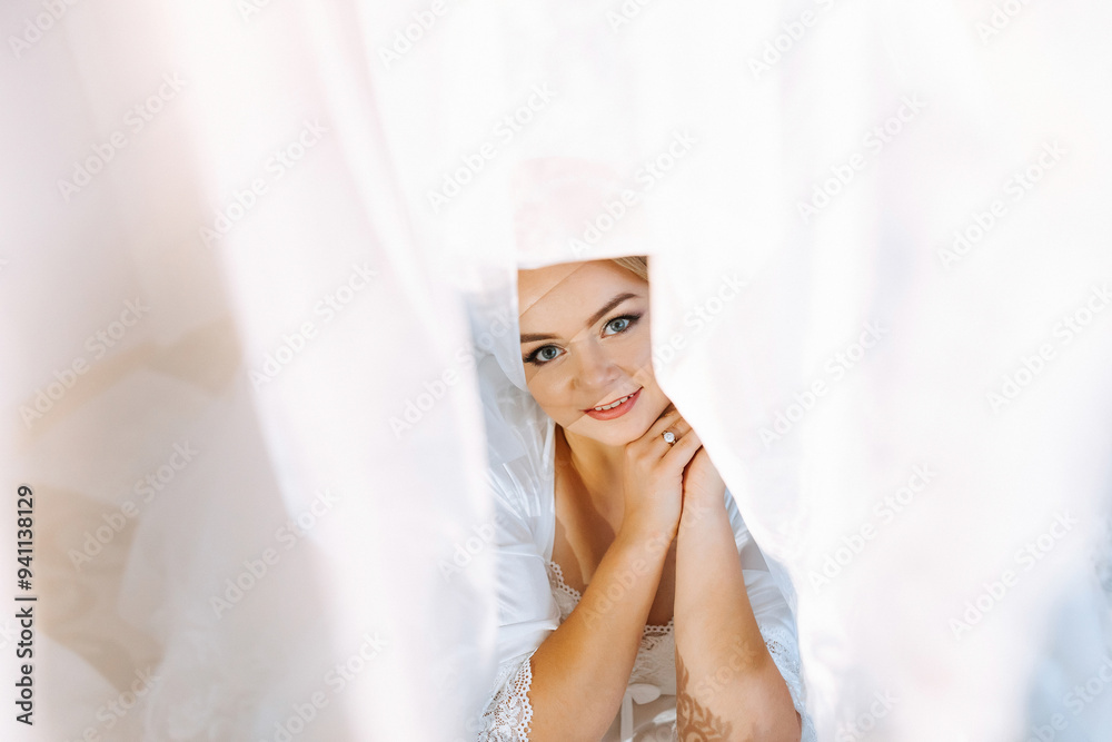 Fototapeta premium A woman is standing behind a curtain, with her face partially hidden. She is wearing a white dress and has her hands on her hips. Concept of mystery and intrigue