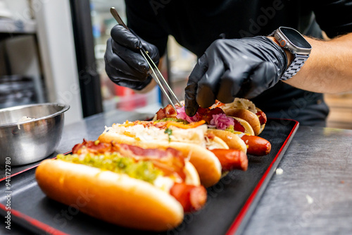 close-up of hands in black gloves using tongs to garnish hot dogs on black tray. scene captures the preparation of gourmet street food, emphasizing the attention to detail and culinary craftsmanship. photo