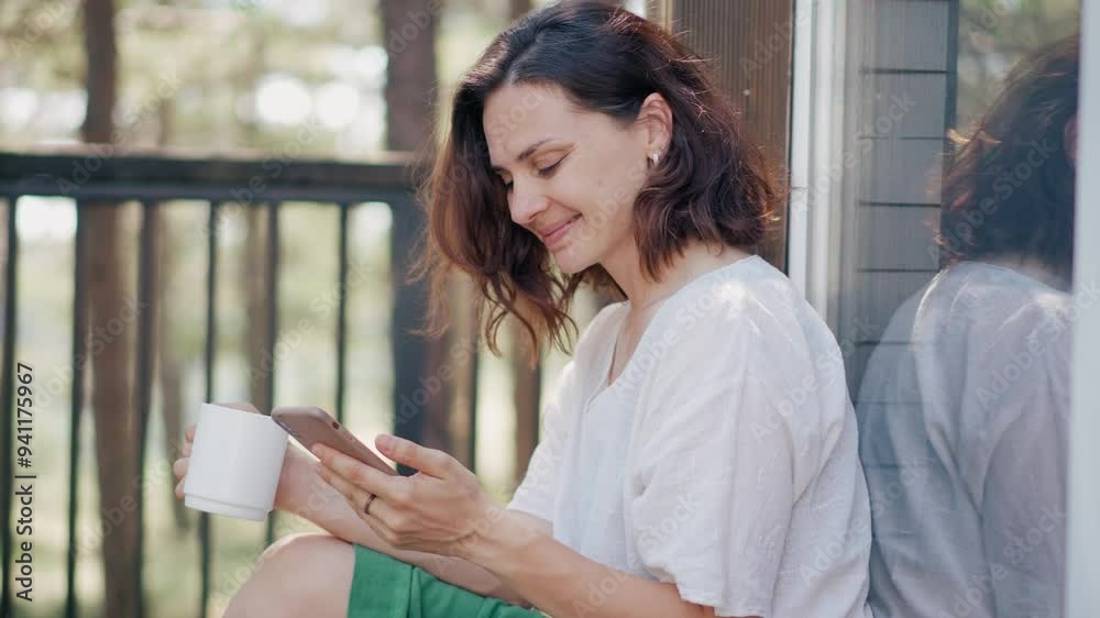 A young Caucasian woman using her smartphone while sitting on the veranda of a wooden country house in the woods