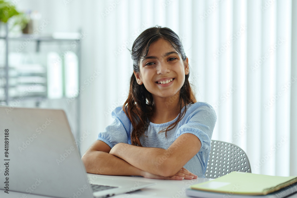 Portrait of smiling young girl with crossed arms sitting at desk with laptop opposite window shades showing white teeth and wearing blue shirt Portrait of smiling young girl with crossed arms sitting at desk with laptop opposite window shades showing white teeth and wearing blue shirt