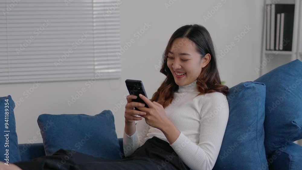 Young woman is using her smartphone while relaxing on a blue sofa at home. She is smiling and seems to be enjoying herself Young woman is using her smartphone while relaxing on a blue sofa at home. She is smiling and seems to be enjoying herself