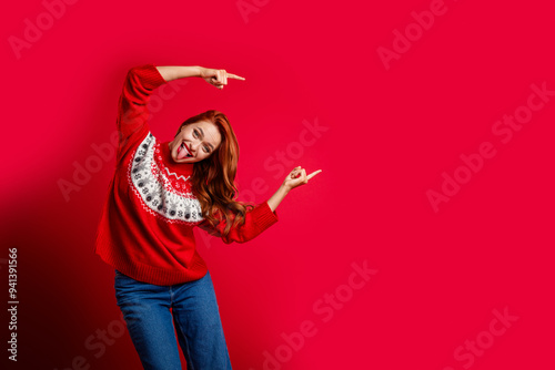 Photo of redhair young attractive funn girl tongue out wearing ugly sweater xmas style pointing fingers present isolated on red color background photo