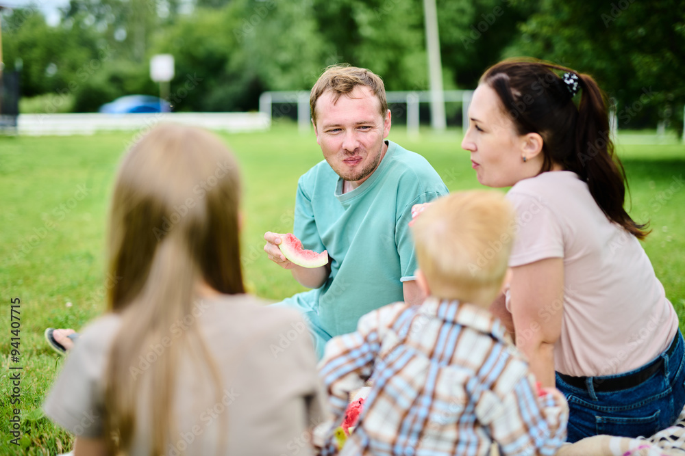 Happy Caucasian family on picnic. Mom, dad, son and daughter sitting on picnic blanket on green grass in yard, eating watermelon and having fun Happy Caucasian family on picnic. Mom, dad, son and daughter sitting on picnic blanket on green grass in yard, eating watermelon and having fun