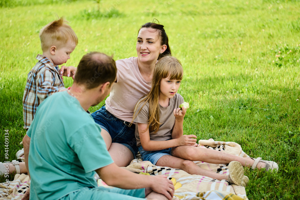 Happy Caucasian family on picnic. Mom, dad, son and daughter sitting on picnic blanket on green grass in yard, eating watermelon and having fun Happy Caucasian family on picnic. Mom, dad, son and daughter sitting on picnic blanket on green grass in yard, eating watermelon and having fun