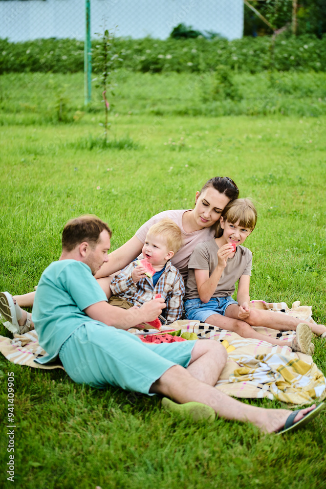 Happy Caucasian family on picnic. Mom, dad, son and daughter sitting on picnic blanket on green grass in yard, eating watermelon and having fun Happy Caucasian family on picnic. Mom, dad, son and daughter sitting on picnic blanket on green grass in yard, eating watermelon and having fun