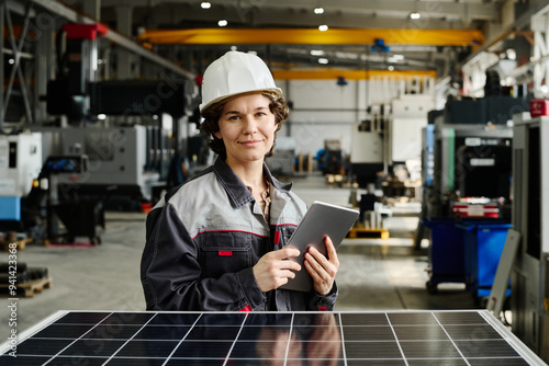 Portrait of female engineer standing in large industrial workshop holding tablet while smiling at camera photo