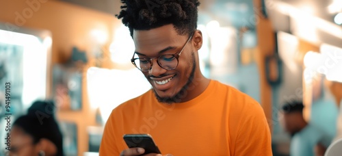 African Americans Gen X Millenial black male all smiles in an orange shirt and glasses focused on phone as he scrolls through mobile cell with black hair and blurred background 