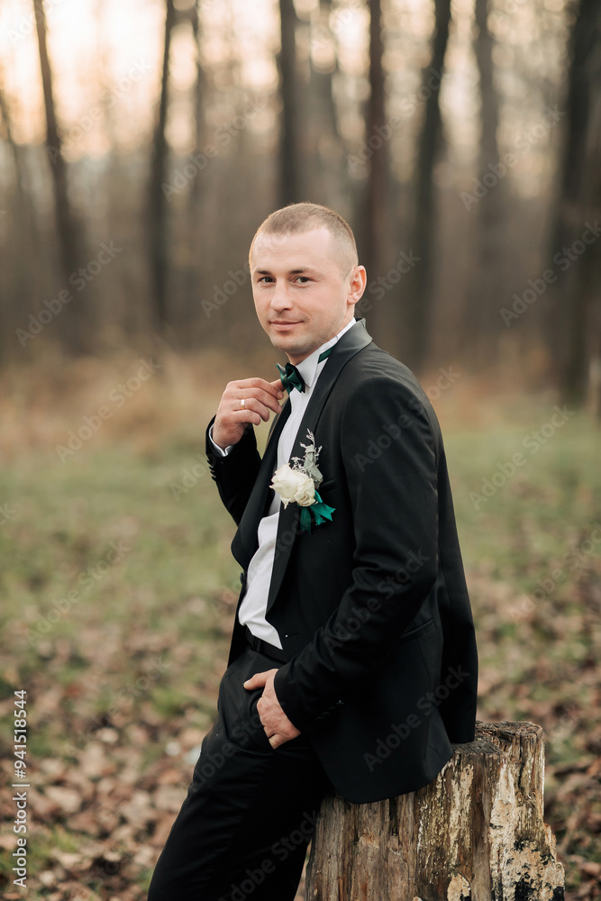 A man in a black suit and white shirt is posing for a picture in a forest. He is wearing a green flower in his lapel and a bow tie. Scene is elegant and romantic A man in a black suit and white shirt is posing for a picture in a forest. He is wearing a green flower in his lapel and a bow tie. Scene is elegant and romantic