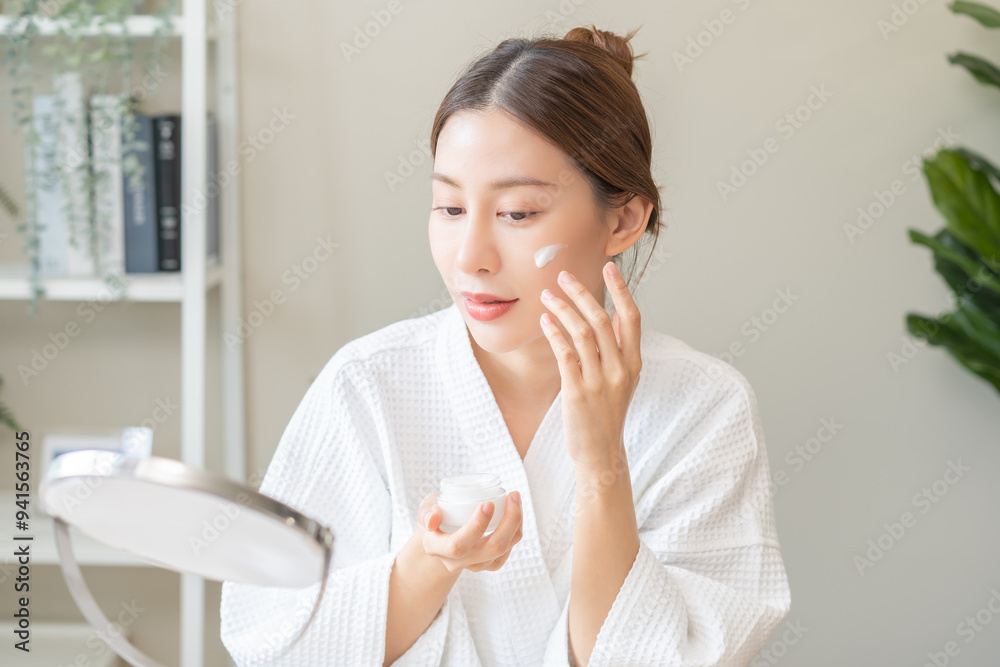 Facial beauty skin care, smile of pretty asian young woman in bathrobe looking at mirror, hand applying moisturizer lotion on her face, putting cream treatment before makeup cosmetic routine at home. Facial beauty skin care, smile of pretty asian young woman in bathrobe looking at mirror, hand applying moisturizer lotion on her face, putting cream treatment before makeup cosmetic routine at home.