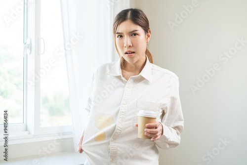 Cloth stain, disappointment asian young woman clumsy with hot coffee, tea stains on shirt, hand show making spill drop on white t-shirt, spot dirty or smudge on clothes at home, isolated on background photo