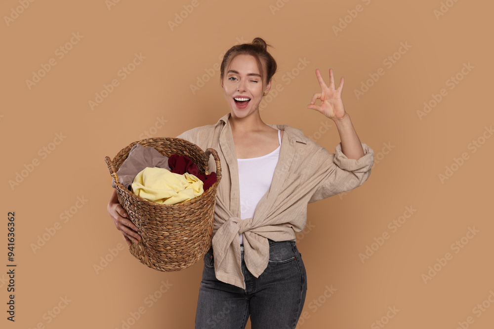 Happy young housewife with basket full of laundry showing OK gesture on pale orange background Happy young housewife with basket full of laundry showing OK gesture on pale orange background
